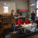 Cztery osoby są w bibliotece na wydarzeniu; jedna kobieta stoi z mikrofonem i mówi, podczas gdy trzech mężczyzn stoi obok niej. Za nimi jest ekran pokazujący okładkę książki, a regały wypełniają tło.