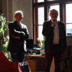 Dwóch starszych mężczyzn stoi w oświetlonej słońcem bibliotece; jeden trzyma mikrofon i mówi, podczas gdy drugi stoi ze skrzyżowanymi ramionami i się uśmiecha. Tło wypełniają regały z książkami i duże okna, przez które wpada światło.