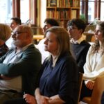 Zróżnicowana grupa ludzi siedzi w rzędach krzeseł w bibliotece lub księgarni, uważnie słuchając prezentacji lub wydarzenia, w tle widać regały z książkami i duże okna.