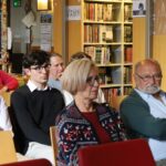 Grupa ludzi, zarówno mężczyzn, jak i kobiet w różnym wieku, siedzi uważnie w bibliotece lub księgarni, zwrócona twarzą do przodu. W tle widoczne są regały wypełnione książkami.