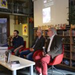 Trzech mężczyzn siedzi na czerwonych krzesłach, mówiąc do mikrofonów na wydarzeniu książkowym w bibliotece. Na ścianach ustawione są regały z książkami i plakaty, a ekran projekcyjny wyświetla okładkę książki za nimi. Stół z książkami i wodą znajduje się z przodu.