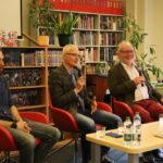 Trzech mężczyzn siedzi na czerwonych krzesłach, trzymając mikrofony i przemawia na wydarzeniu w bibliotece lub księgarni. W tle widać regały na książki, zielone rośliny i stół z butelkami z wodą i książkami.