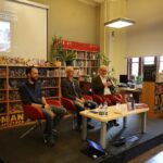 Trzech mężczyzn siedzi na małej scenie w bibliotece, zaangażowanych w dyskusję lub wydarzenie książkowe. Za nimi znajdują się półki wypełnione książkami i roślinami, a ekran projekcyjny wyświetla obraz góry i tekst w języku angielskim.