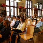 Grupa osób siedzi na drewnianych krzesłach w bibliotece lub klasie, patrząc przed siebie i uważnie słuchając. W tle widoczne są regały na książki i duże okna.