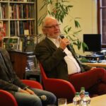 Dwóch starszych mężczyzn siedzi na czerwonych krzesłach podczas dyskusji w bibliotece. Jeden mężczyzna, trzymający mikrofon, mówi, a drugi słucha. W tle widać regały z książkami, rośliny i okno.