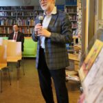 Mężczyzna o siwych włosach i okularach mówi do mikrofonu, stojąc w bibliotece; w tle siedzi uśmiechnięty mężczyzna w garniturze, skąd widać regały z książkami i krzesła.