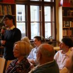 Młody mężczyzna stoi, trzyma mikrofon i przemawia do uważnej publiczności siedzącej w przytulnej bibliotece pełnej regałów z książkami i dużych okien.