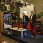 Cztery osoby stoją z przodu sali bibliotecznej przy ekranie i czerwonych krzesłach, z książkami i mikrofonem na stole. Ściany są ustawione wzdłuż regałów, a po lewej stronie widoczny jest baner o bibliotece uniwersyteckiej.