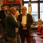 Dwóch starszych mężczyzn stoi i rozmawia w bibliotece lub księgarni, podczas gdy inni rozmawiają w tle. W ciepło oświetlonym pomieszczeniu widać regały na książki, czerwone krzesła i duże okna.