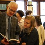 Mężczyzna w garniturze i krawacie pokazuje książkę kobiecie o długich blond włosach w zatłoczonej księgarni lub bibliotece, w tle znajduje się kilka innych osób przeglądających książki.