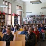 Grupa ludzi siedzi na drewnianych krzesłach w bibliotece, uważnie patrząc przed siebie. Tło wypełniają regały z książkami, a duże okna wpuszczają naturalne światło. Zegar i kilka prostokątnych lamp zwisa z sufitu.