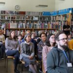 Grupa osób siedzi uważnie w bibliotece, zwrócona twarzą do przodu. Ściany są wyłożone regałami wypełnionymi książkami, a nad półkami widoczny jest zegar. Jedna osoba stoi z tyłu pomieszczenia.