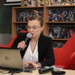 Kobieta w okularach i czarnym blezerze mówi do mikrofonu, siedząc przy stole z laptopem przed sobą. Znajduje się w pokoju z czerwonymi krzesłami i półkami wypełnionymi książkami i kolorowymi obrazami.