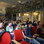 Grupa osób, składająca się z dorosłych i dzieci, siedzi uważnie na krzesłach w bibliotece z wysokimi regałami na książki i antresolą, uczestnicząc w wydarzeniu lub prezentacji.