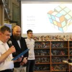 Trzech mężczyzn stoi przed regałem z książkami i ekranem projektora wyświetlającym diagram kostki Rubika 3D i tytuł Kostka Rubika. Jeden mężczyzna mówi do mikrofonu, trzymając dokumenty.