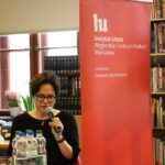 Kobieta o krótkich, ciemnych włosach i w okularach mówi do mikrofonu przy stole z butelkami wody, siedząc obok czerwonego baneru Instytutu Liszta w bibliotece lub księgarni.