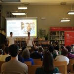 Grupa ludzi uczestniczy w prezentacji w bibliotece. Dwóch mówców stoi z przodu przy wyświetlanym slajdzie ze zdjęciem mężczyzny, podczas gdy publiczność stoi naprzeciwko nich, siedząc w rzędach krzeseł. Półki z książkami otaczają salę.