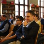 Grupa ludzi siedzi uważnie w bibliotece, patrząc przed siebie. Jeden mężczyzna na pierwszym planie patrzy w kamerę, uśmiecha się i pozuje z ręką na brodzie. W tle widać regały z książkami i duże okna.
