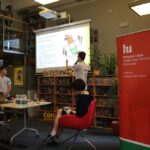 Trzy osoby są w bibliotece podczas prezentacji. Jedna stoi z mikrofonem skierowanym na ekran, druga stoi przy stole z butelkami wody, a trzecia siedzi. Duży czerwony baner znajduje się po prawej stronie.
