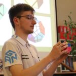 Młody mężczyzna w okularach i białej koszuli trzyma skomplikowaną, wielokolorową kostkę układanki. Za nim projekcja pokazuje obrazy podobnych puzzli. W tle widoczne są półki z książkami, czerwonymi segregatorami i rośliną doniczkową.