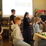Grupa dzieci i dorosłych, w tym zakonnica, stoi w bibliotece przy stole z kolorowymi kostkami Rubika. Ekran projektora w tle wyświetla logo Rubika i polski tekst.