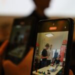 Osoba trzyma smartfon, nagrywając lub przesyłając strumieniowo obraz trzech osób stojących i siedzących przy stole w pomieszczeniu z regałami na książki i sprzętem elektronicznym, podczas gdy w tle widać jeszcze jedną osobę.