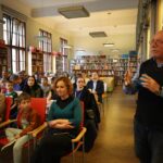 Mężczyzna stoi i przemawia do publiczności siedzącej w bibliotece, z regałami na książki i dużymi oknami w tle. Dorośli i dzieci słuchają uważnie, niektórzy się uśmiechają, siedząc w rzędach czerwonych krzeseł.