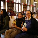 Uśmiechnięta dziewczynka i zakonnica siedzą razem w zatłoczonej bibliotece, trzymając kostki Rubika, a wokół nich siedzą inni dorośli i dzieci. W tle widać regały z książkami i duże okna.