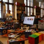 W bibliotece lub klasie rozstawiono stół pokryty różnymi kostkami Rubika i puzzlami, w tle znajdowały się półki z książkami, a w pobliżu siedziało lub stało kilka osób.