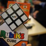 Osoba trzyma kostkę Rubika w pudełku oraz ułożoną kostkę Rubika z samymi białymi polami, podczas gdy w tle na stole rozmazują się postacie ludzkie i pudełka z grą.