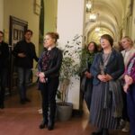 Grupa ludzi stoi uważnie w korytarzu z łukowatymi sufitami i wiszącymi światłami. Większość zwrócona jest do przodu, niektórzy ze skrzyżowanymi ramionami, słuchając czegoś niewidzialnego. W centrum znajduje się doniczkowa roślina.