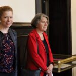 Dwie kobiety stoją w pomieszczeniu przy gablocie. Jedna z rudymi włosami związanymi w kucyk, uśmiecha się i nosi czarną kurtkę; druga ma krótkie siwe włosy i nosi czerwony żakiet, patrząc przed siebie. Sceneria wydaje się być formalna lub instytucjonalna.