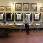 Kobieta stoi przy mikrofonie w galerii sztuki, a za nią na ścianach wiszą oprawione rysunki. Po lewej stronie znajduje się okrągły stół z kwiatami i poczęstunkiem, a nad dziełami sztuki wiszą historyczne zdjęcia.