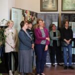 Grupa kobiet stoi uważnie w pokoju, niektóre ze skrzyżowanymi ramionami, przed wyświetlaczami informacyjnymi i oprawionymi zdjęciami na ścianie. Na pierwszym planie widoczna jest roślina doniczkowa.
