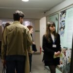 Ludzie oglądają i omawiają duże plakaty z ilustracjami i tekstem, zawieszone na ścianach korytarza; jedna kobieta stoi przy plakatcie, ma na sobie identyfikator i trzyma papiery.