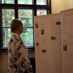 Osoba ubrana w białą i niebieską koszulę z kwiatowym wzorem stoi wewnątrz budynku i czyta informację o Stefanie Zweigu, a za oknem widać duże okna i zielone drzewa.