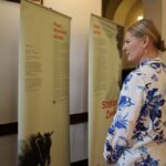 Kobieta w białej bluzce z niebieskimi wzorami kwiatowymi stoi w środku, czytając panele wystawy z tekstem i obrazami. Na banerach znajdują się informacje w języku polskim i niemieckim, a jeden z nich przedstawia część pyska krowy.