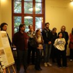 Grupa ludzi stoi uważnie w pokoju z beżowymi ścianami i dużym oknem, patrząc na coś poza kadrem. Tablica informacyjna jest widoczna po lewej stronie obrazu.