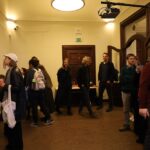 Grupa ludzi w ciepło oświetlonym pomieszczeniu, niektórzy patrzą na dzieła sztuki na ścianie, podczas gdy inni stoją i rozmawiają. Na suficie zamontowany jest projektor, a w tle widać drzwi.