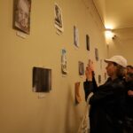 Osoba w białej czapce i czarnej kurtce robi zdjęcie dzieł sztuki wystawionych na beżowej ścianie w galerii sztuki, podczas gdy inni zwiedzający oglądają eksponaty.