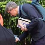 Osoba o siwych włosach, w okularach i z plecakiem, pochyla się, trzyma książki i przegląda zawartość pudełka znajdującego się na zewnątrz, w pobliżu zielonych krzewów.