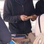 Mężczyzna w okularach i ciemnej bluzie z kapturem ogląda książkę na targu książek na świeżym powietrzu. Przed nim stoi pudełko pełne różnorodnych książek, a w pobliżu znajdują się inni ludzie przeglądający książki.