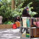 Osoba przegląda książki umieszczone na wózku z napisem Kiermasz Książki na plenerowych targach książki, podczas gdy inni ludzie siedzą i rozmawiają na zacienionym dziedzińcu otoczonym zielenią.