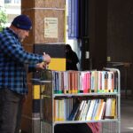 Mężczyzna w niebieskiej kraciastej koszuli i czapce układa książki na wózku na zewnątrz, w pobliżu budynku. Na słupie widnieje napis Kiermasz książki, oznaczający targi książki. W tle widać drzewa i ludzi.