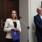 Kobieta w kremowej marynarce przemawia do mikrofonu, trzymając teczkę, podczas gdy w pobliżu stoi mężczyzna w ciemnym garniturze. Oboje znajdują się w środku, w tle widać oprawione obrazy i drewniane drzwi.