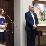 Kobieta stoi przy mikrofonie trzymając folder, podczas gdy mężczyzna w garniturze stoi w pobliżu. Oboje są w środku, obok ekspozycji z dziełami sztuki architektonicznej i tekstem.