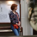 Kobieta z kręconymi rudymi włosami, w okularach, w kwiecistej koszuli, dżinsach i pomarańczowej torbie na ramię, stoi w pomieszczeniu przed tablicą z napisem Walden. Na pierwszym planie widać rozmazane liście.