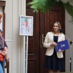 Kobieta w białej marynarce przemawia do mikrofonu, trzymając w ręku niebieską teczkę, podczas gdy inna kobieta z kręconymi, rudymi włosami i pomarańczową torbą słucha jej w pomieszczeniu znajdującym się nieopodal, z drewnianymi drzwiami i oprawionymi obrazami na ścianach.