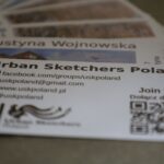 Zbliżenie na wizytówkę Urban Sketchers Poland, na której widać dane kontaktowe, ikony mediów społecznościowych, kod QR i częściowo widoczne wizytówki z nazwiskiem Justyna Wojnowska.