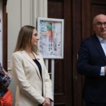 Trzech dorosłych stoi w pomieszczeniu przy mikrofonie; mężczyzna w ciemnym garniturze mówi, podczas gdy dwie kobiety, jedna w białym blezerze, a druga we wzorzystej koszuli z pomarańczową torbą, słuchają. W tle wyświetlana jest oprawiona praca artystyczna.